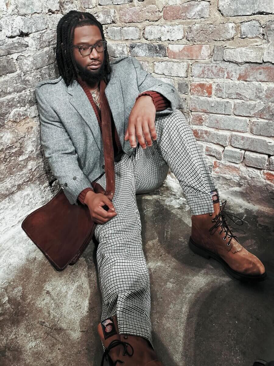 She Curates One of Birmingham’s Top Vintage & Consignment Boutiques - 6 A man wearing a gray blazer, patterned pants, and brown boots sits on the floor against a brick wall, holding a brown leather bag from a Birmingham vintage boutique.