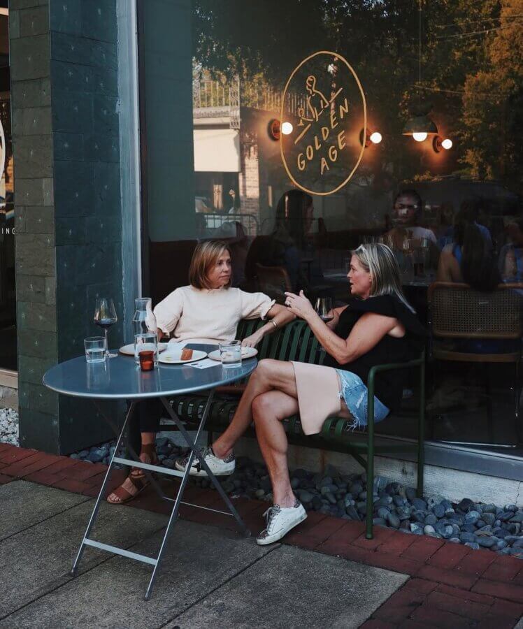 Your Guide to Mountain Brook's Villages - 4 Two women sit and talk at an outdoor café table with drinks, in front of a window labeled "Golden Age" in Mountain Brook. Other patrons are visible inside, capturing a lively scene for the Villages Guide.