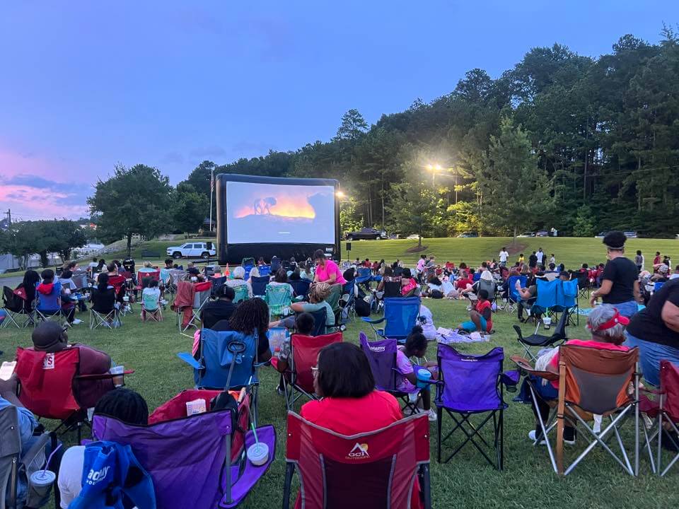 A crowd of people sits on lawn chairs and blankets watching an outdoor movie on a large screen in a park at dusk, making it the perfect spot if you're searching for where to watch Birmingham outdoor movies.