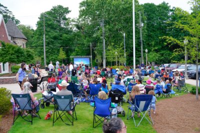 A large group of people in lawn chairs enjoy a Birmingham outdoor movie screening on a grassy area near a parking lot and trees.