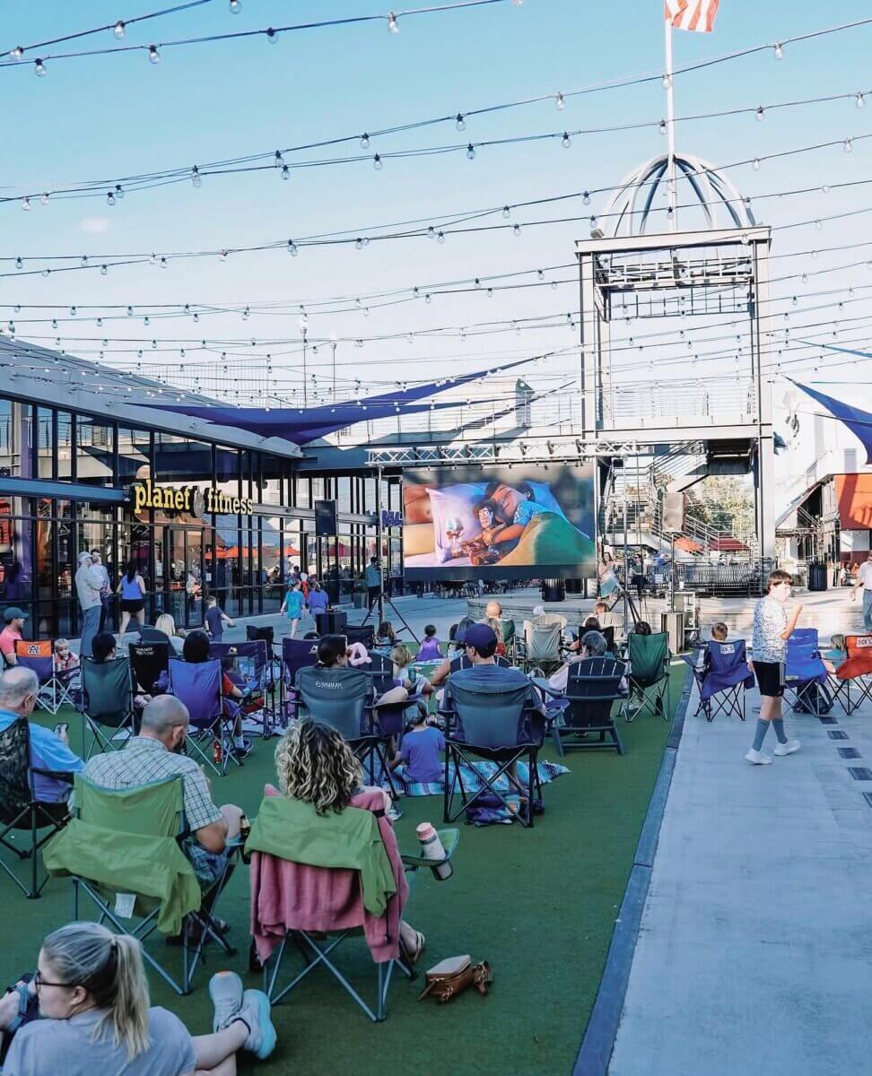 People sit in folding chairs on artificial grass, watching an outdoor movie in Birmingham’s shopping center under string lights and a blue sky—a perfect scene for any movie guide.