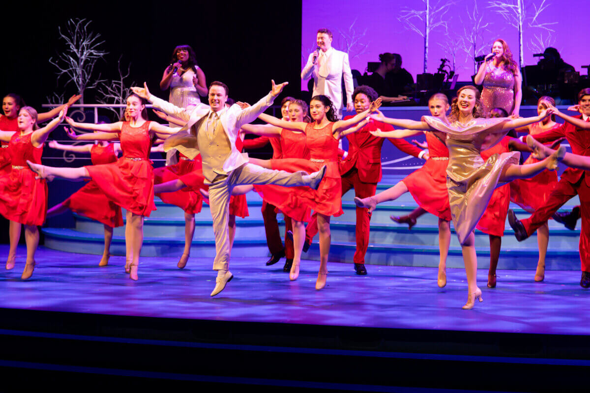 A group of dancers in red dresses and beige suits perform a synchronized kickline on a brightly lit stage at Red Mountain Theater, with singers in the background.
