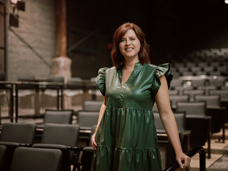 The Birmingham Stage Everyone's Talking About: Inside Red Mountain Theatre - 4 A woman in a green dress stands in an empty auditorium at Red Mountain Theater, smiling at the camera. Rows of gray seats and industrial-style walls at the nonprofit theater.