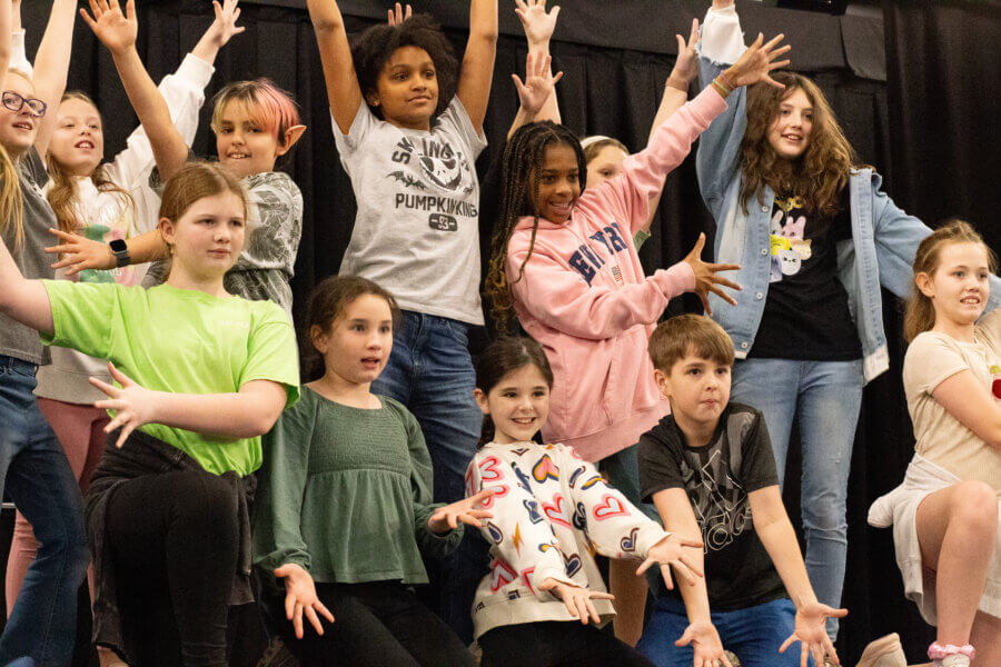 The Birmingham Stage Everyone's Talking About: Inside Red Mountain Theatre - 6 A group of children pose enthusiastically on stage at Red Mountain Theater, with arms outstretched and smiling faces, in front of a black curtain backdrop.