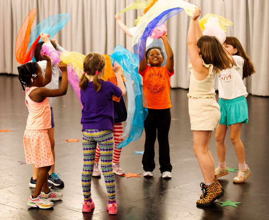 The Birmingham Stage Everyone's Talking About: Inside Red Mountain Theatre - 3 A group of children stand in a circle, holding colorful scarves above their heads in a dance studio at Red Mountain Theater, with a gray curtain in the background.