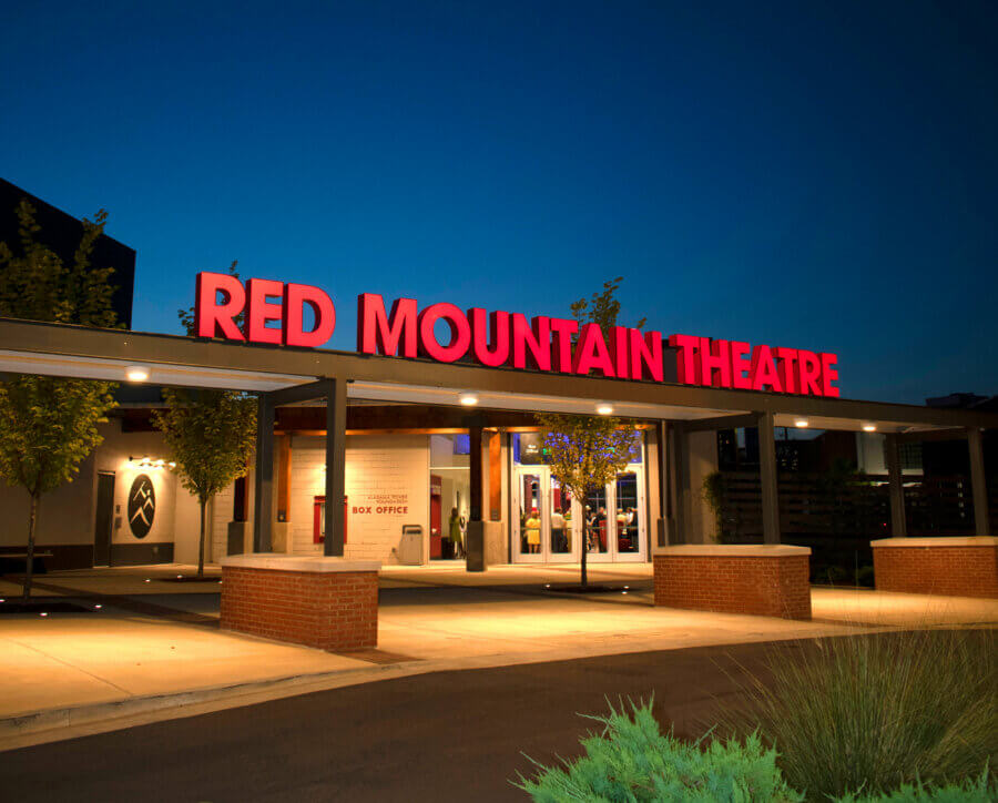 The Birmingham Stage Everyone's Talking About: Inside Red Mountain Theatre - 2 Entrance of Red Mountain Theater at dusk, featuring a large red illuminated sign above the walkway, with trees and the nonprofit theater’s box office visible in the background.