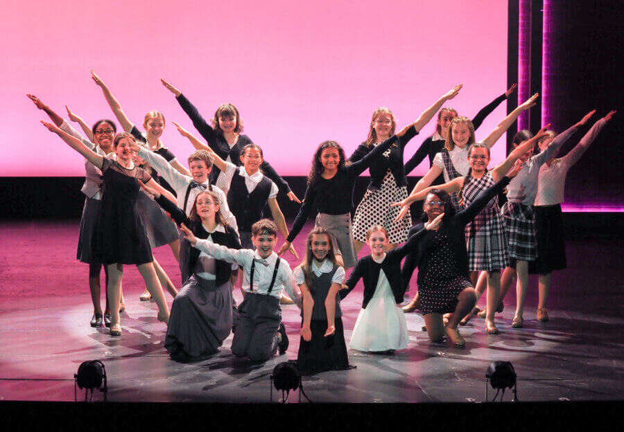 The Birmingham Stage Everyone's Talking About: Inside Red Mountain Theatre - 7 A group of children pose on stage in coordinated outfits, standing and kneeling with arms outstretched against a pink-lit background at Red Mountain Theater.