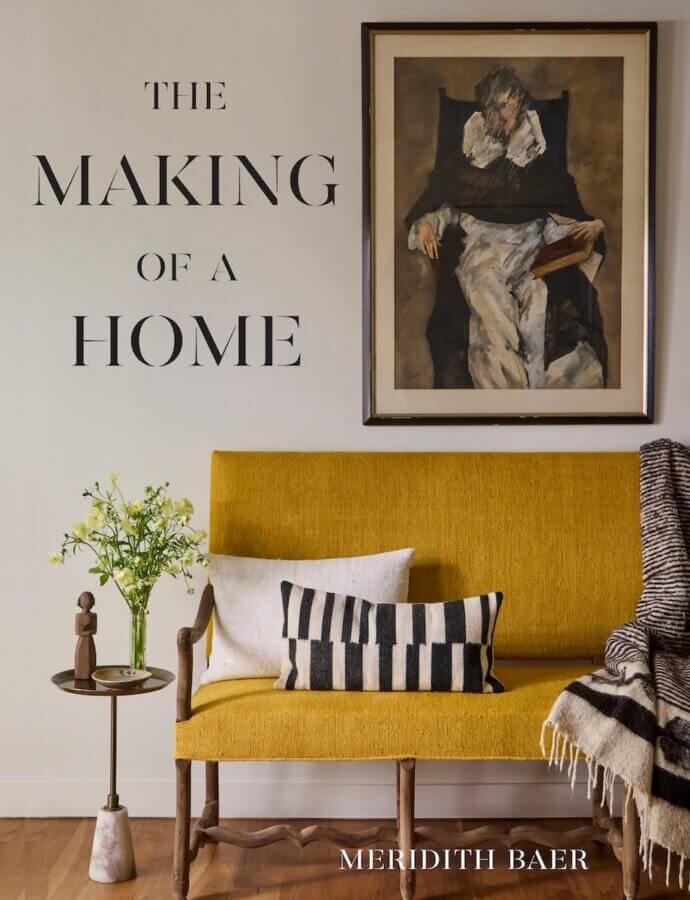 9 New Design-Forward Coffee Table Books for Your Wish List - 10 A mustard yellow bench with cushions and a throw blanket sits below a painting; a small table with flowers and decor is beside it. This scene exudes Design Envy, embodying the style of “The Making of a Home” by Meridith Baer.