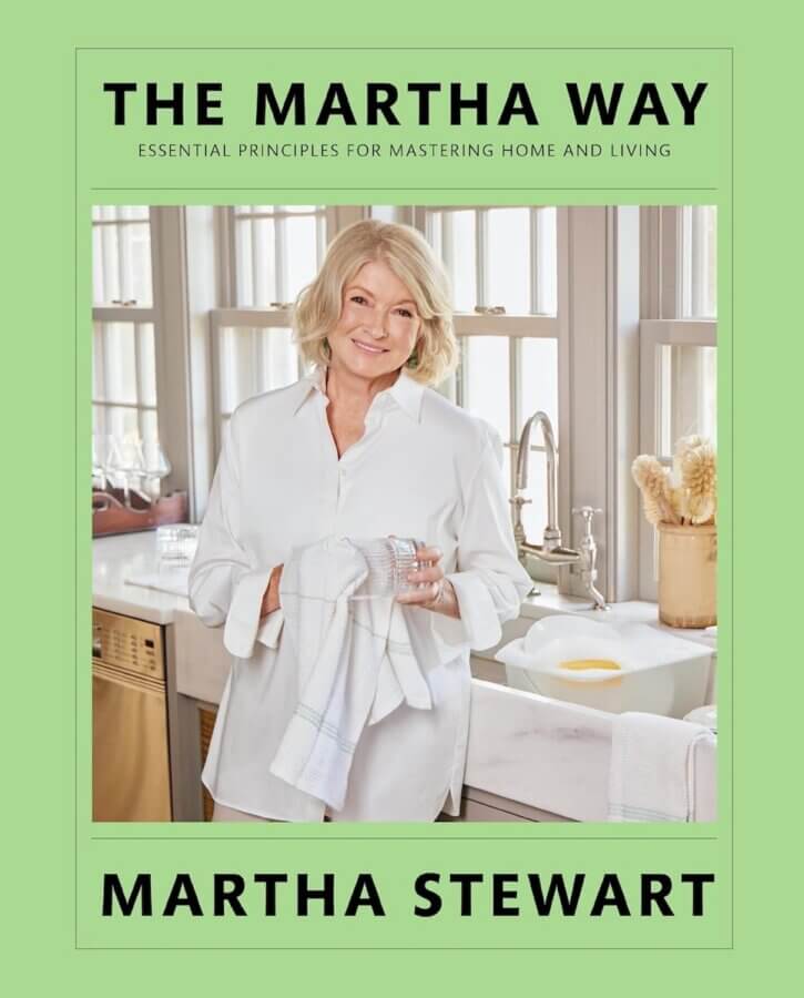 9 New Design-Forward Coffee Table Books for Your Wish List - 8 A woman in a white shirt stands in a kitchen drying a plate with a towel. The image graces the cover of "The Martha Way" by Martha Stewart.