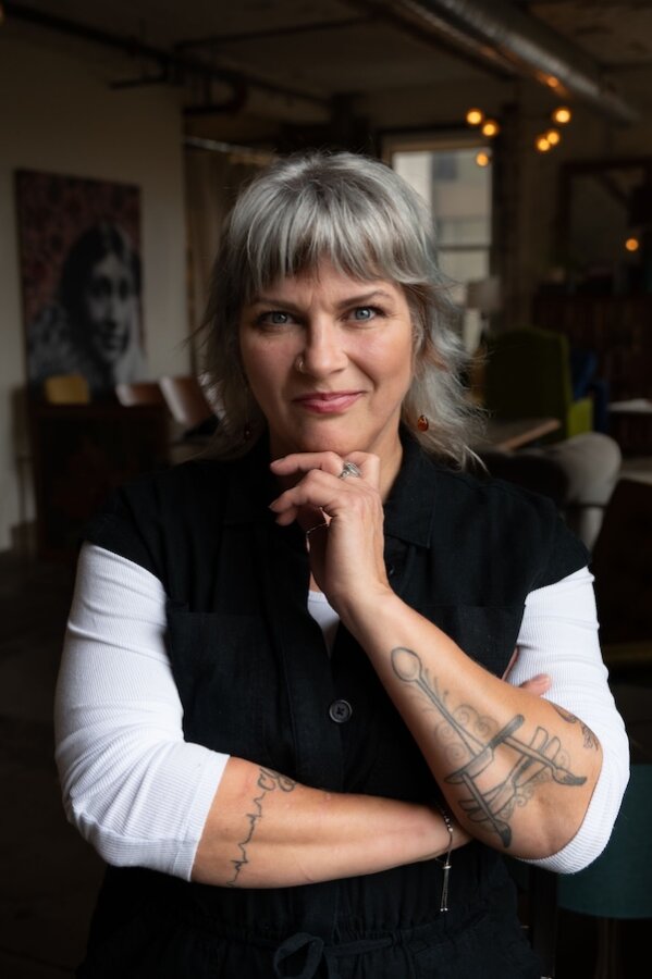 Discover the "Queens of Cuisine," A New Women-Led Culinary Trail in Greenville, SC - 9 A woman with gray hair and tattoos on her forearms stands indoors with her hand resting on her chin, looking at the camera—an inspiring figure among women in the Culinary Trail.