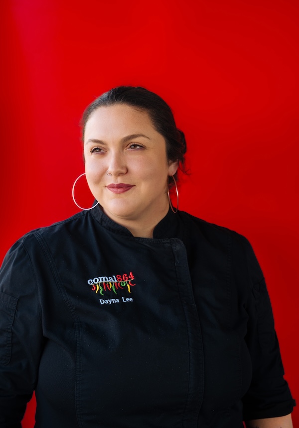 Discover the "Queens of Cuisine," A New Women-Led Culinary Trail in Greenville, SC - 5 A woman representing the Women in Culinary Trail wears large hoop earrings and a black chef's jacket embroidered with "Dayna Lee," standing confidently against a bright red background.