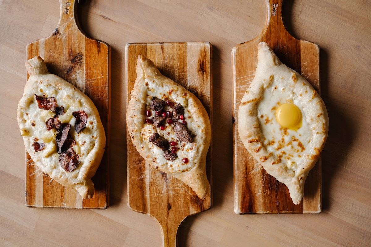 Three khachapuri breads on wooden boards celebrate the Women in Culinary Trail—one topped with bacon, another with meat and pomegranate seeds, and a third featuring a whole egg nestled in melted cheese.