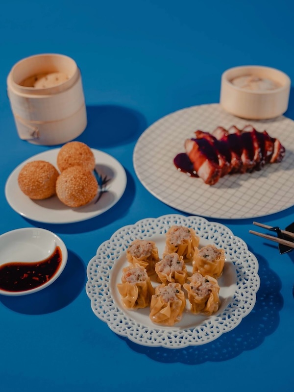 Discover the "Queens of Cuisine," A New Women-Led Culinary Trail in Greenville, SC - 8 A variety of dim sum dishes, inspired by the Women in Culinary Trail, including siu mai, sesame balls, sliced barbecue pork, and steamed buns, are arranged on plates and bamboo steamers on a blue table with a dish of soy sauce.