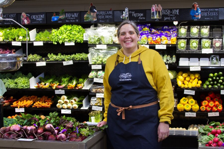 Discover the "Queens of Cuisine," A New Women-Led Culinary Trail in Greenville, SC - 4 A woman from the Women in Culinary Trail, wearing a yellow hoodie and navy apron, stands and smiles in front of a display of fresh vegetables at a grocery store or market.