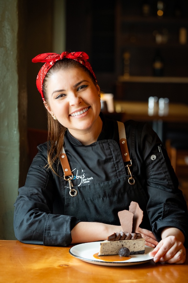 Discover the "Queens of Cuisine," A New Women-Led Culinary Trail in Greenville, SC - 2 A smiling chef, representing the Women in Culinary Trail, sits at a table in a black uniform and red bandana, proudly presenting a plated dessert.