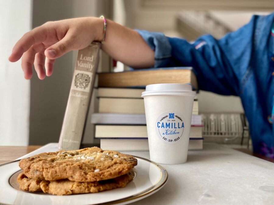 Discover the "Queens of Cuisine," A New Women-Led Culinary Trail in Greenville, SC - 3 A person reaches for a book near a stack of books, a plate of cookies, and a "Camilla Kitchen" coffee cup on a table, celebrating the Women in Culinary Trail.