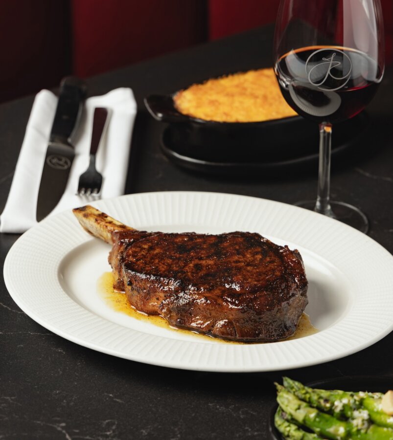 All-Ages Adventure: The Ultimate Trip to Louisville, KY - 18 A grilled bone-in steak on a white plate, served with a side dish and a glass of red wine—an indulgent dining experience that's one of the top things to do in Louisville, complete with knife and fork on a napkin in the background.