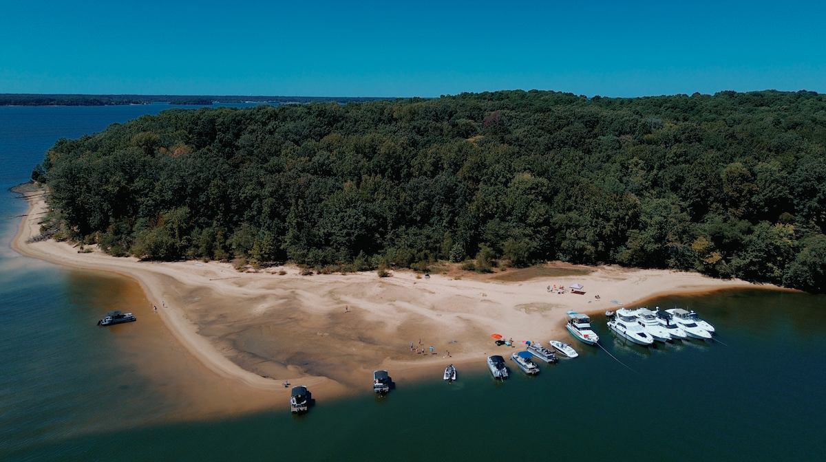 6 Southern Waterfront Weekend Escapes - 12 Aerial view of a sandy beach with several boats docked along the shore and a forested area in the background, perfect for relaxing waterfront vacations on a sunny day.