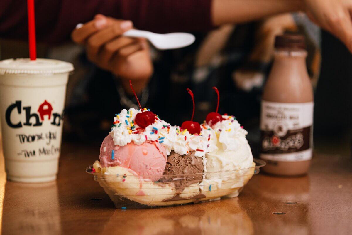 A banana split with scoops of strawberry, chocolate, and vanilla ice cream, topped with whipped cream, cherries, and sprinkles sits next to a milkshake and chocolate milk bottle—perfect for a Southern family vacation or fun family travel treat.