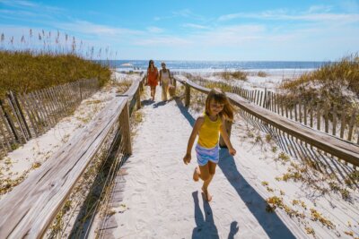 A young girl runs along a sandy path to the beach during family vacations, while two adults walk behind her on a sunny day with clear skies and the ocean in the background.