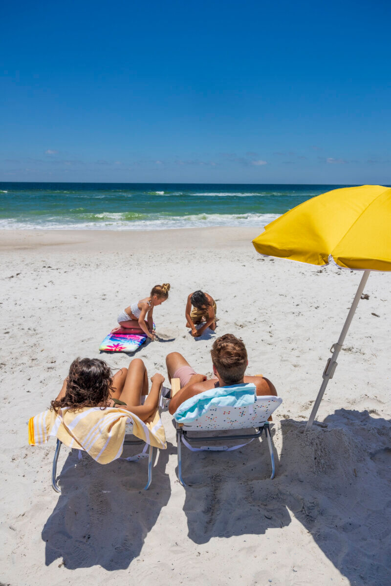 Two adults sit on beach chairs under a yellow umbrella while two children play on a towel nearby, all facing the ocean—a perfect scene for a kid-tested, parent-approved Southern family vacation on a sunny day.