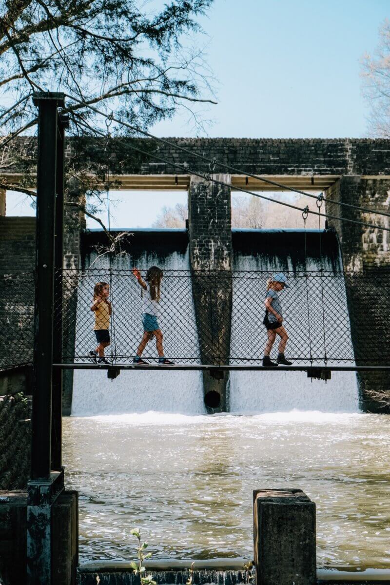 Three children walk across a suspended rope bridge in front of a dam with water flowing below—a perfect scene for parent-approved, kid-tested Southern family vacation destinations.