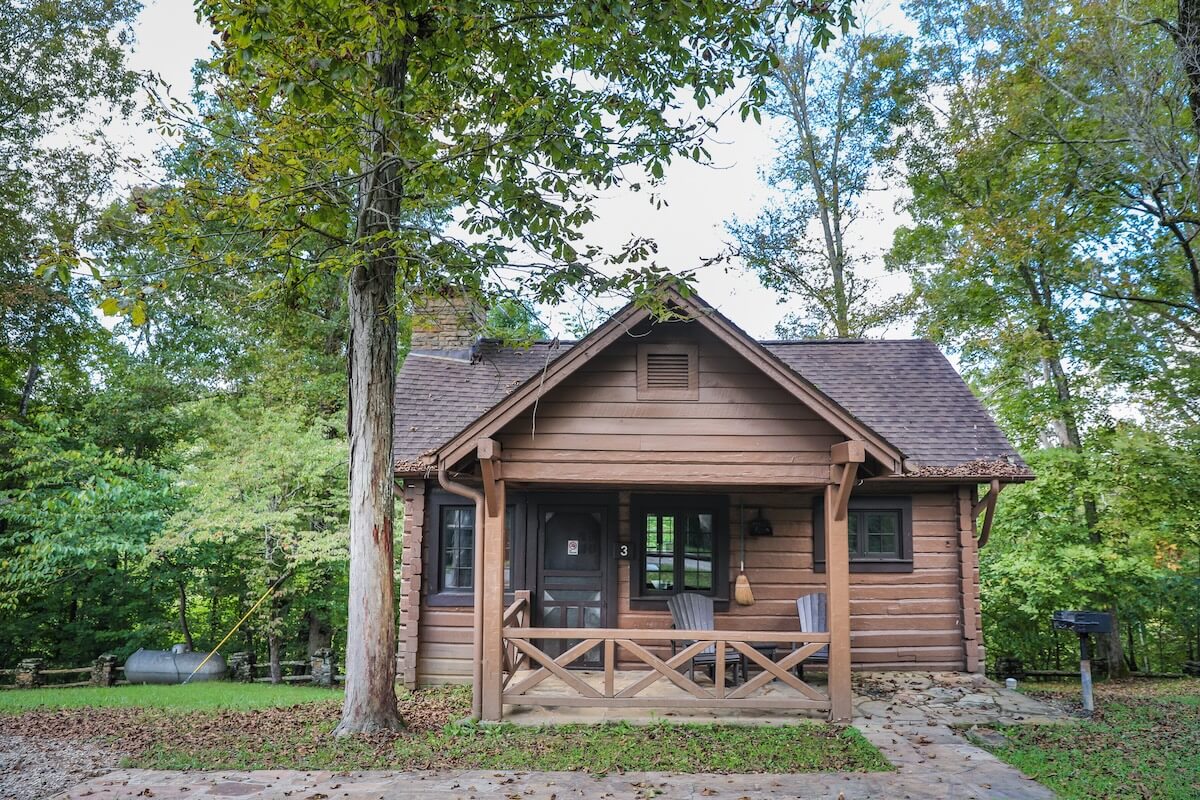 A small wooden log cabin with a front porch, two chairs, and a railing, surrounded by trees and greenery—perfect for relaxing family vacations.