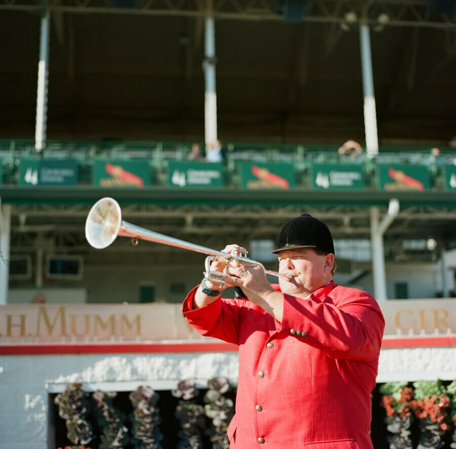 Your Derby 2026 Playbook: What to Know Before You Go - 3 A man in a red coat and black hat plays a silver trumpet outside at the Kentucky Derby, with stadium seating and a green sign visible in the background.