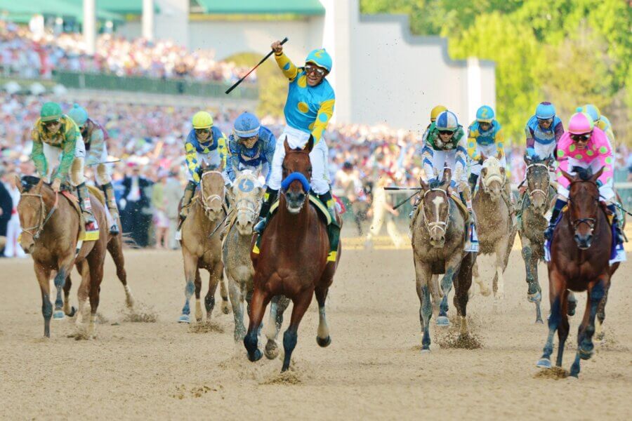 Your Derby 2026 Playbook: What to Know Before You Go - 4 A group of jockeys ride their horses on a dirt racetrack during the Kentucky Derby, with one horse in the lead as the crowd watches in the background.