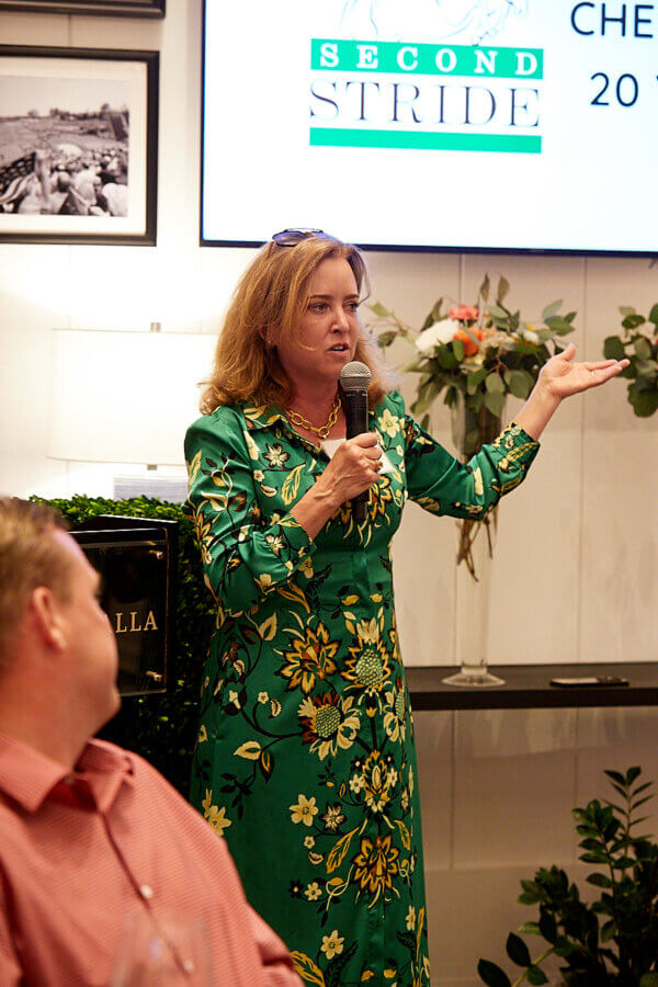 Meet the Louisville Woman Giving Retired Racehorses a Second Chance - 5 Kim Smith, wearing a green floral dress, speaks into a microphone at an indoor event, with a "Second Stride" sign in the background.