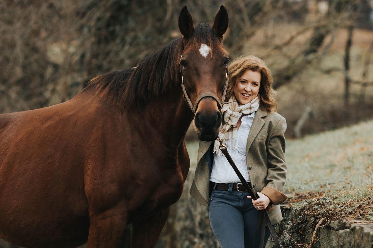 Kim Smith stands outdoors in a blazer and scarf, holding the reins of a brown retired racing Thoroughbred beside her on a grassy path, showcasing the horse’s inspiring second act.