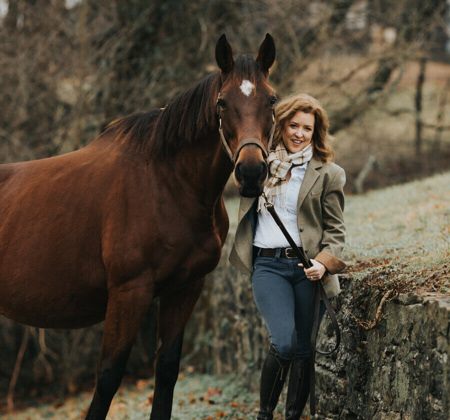 Meet the Louisville Woman Giving Retired Racehorses a Second Chance - 3 Kim Smith, in riding attire, stands beside a retired racing thoroughbred on a grassy, stone-lined path with trees in the background.