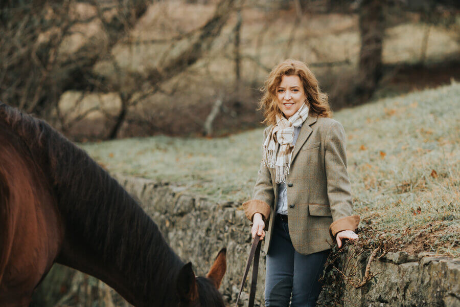 Meet the Louisville Woman Giving Retired Racehorses a Second Chance - 2 A woman in a tweed jacket and scarf stands outdoors holding the reins of a brown horse, with a grassy and wooded background.