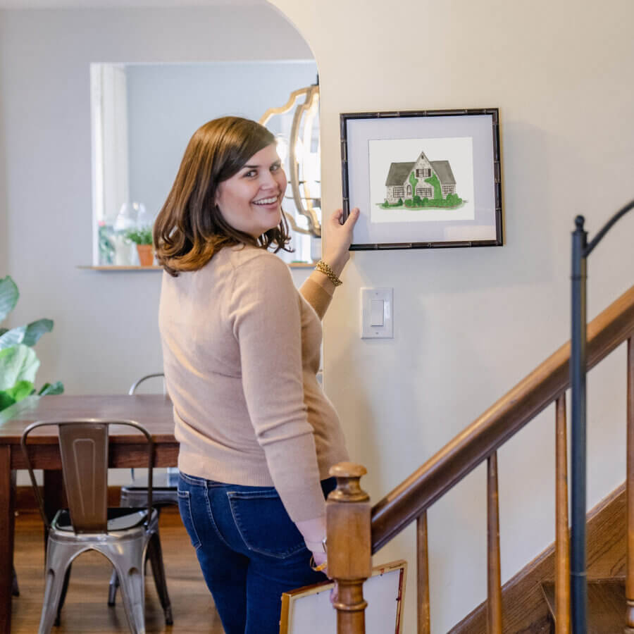 Meet Louisville Artist Maizie Clarke - 2 A woman stands by a staircase, smiling at the camera while hanging a framed illustration of a house on the wall.