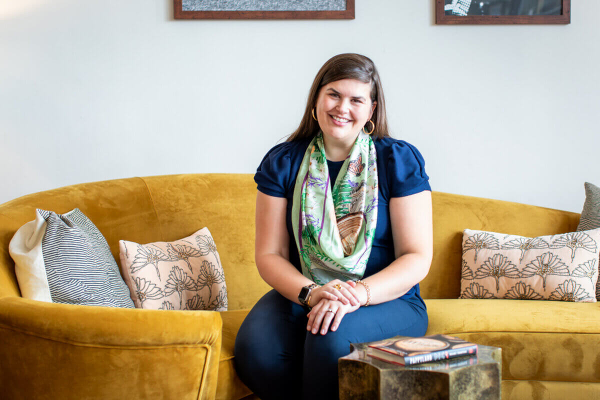 Maizie Clark, known for her creative business and charming illustrations, sits on a yellow couch, smiling, wearing a dark blue top and green scarf. Patterned pillows and books rest on the table in front of her.