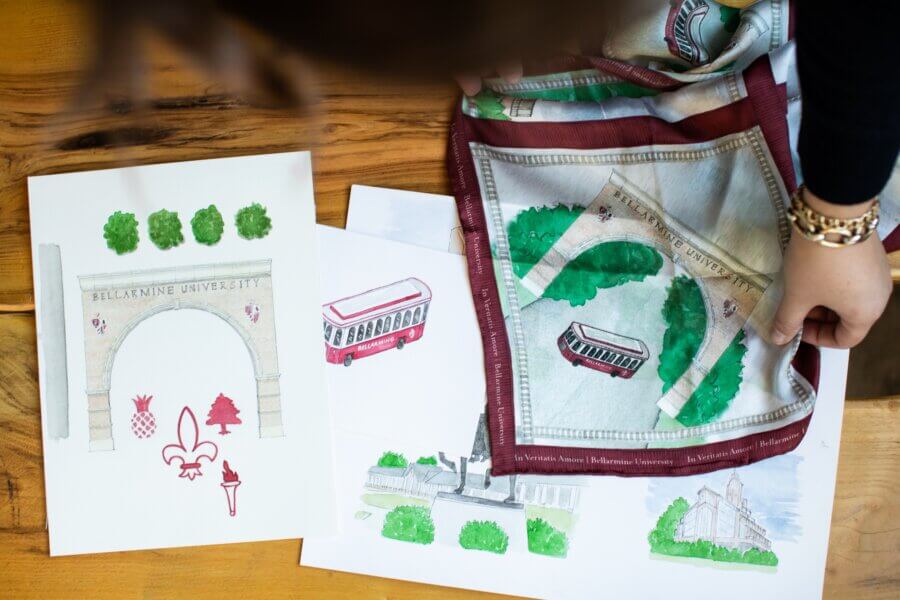 Meet Louisville Artist Maizie Clarke - 4 A person arranges Maizie Clark illustrations and a scarf featuring Bellarmine University landmarks—including an archway, bus, and statue—on a wooden table.