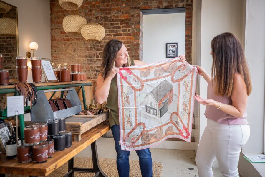 Meet Louisville Artist Maizie Clarke - 6 Two women stand in a shop, holding up and examining a square scarf featuring Maizie Clark's charming house illustrations, next to a display of leather goods and accessories.