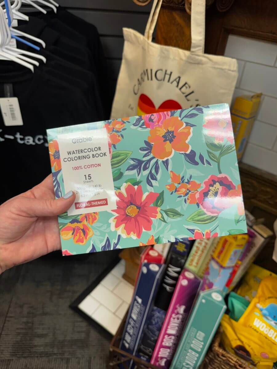 15 Louisville FINDS for May: Spring Fashion, Entertaining Essentials & More - 13 A hand holds a floral-themed watercolor coloring book in a Louisville shop, with T-shirts, a tote bag, and board games among the May FINDS visible in the background.