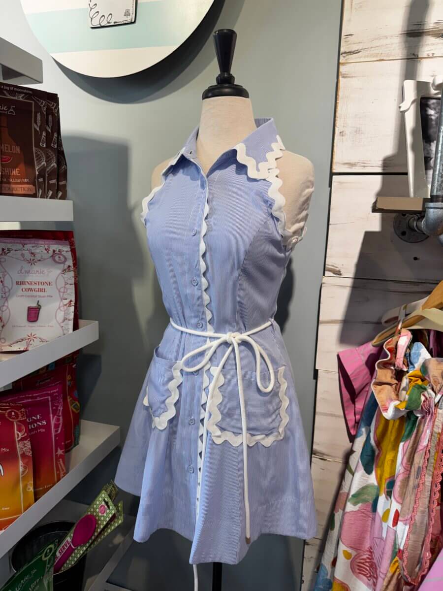 15 Louisville FINDS for May: Spring Fashion, Entertaining Essentials & More - 6 A dress form in Louisville FINDS displays a sleeveless, light blue button-down dress with white scalloped trim and a waist tie, perfect for May, set in a boutique with various packaged goods nearby.