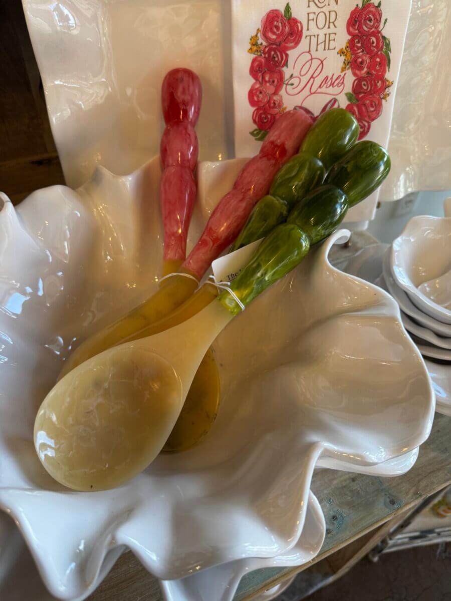 15 Louisville FINDS for May: Spring Fashion, Entertaining Essentials & More - 7 Ceramic serving utensils with pink and green handles rest in a wavy white ceramic bowl, next to stacked plates and a decorative rose napkinβa charming May FINDS highlight for your Louisville table setting.