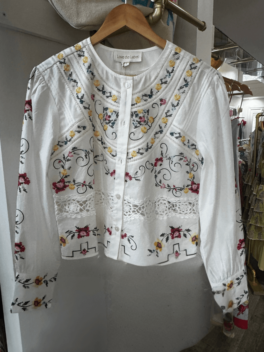 15 Louisville FINDS for May: Spring Fashion, Entertaining Essentials & More - 2 A white long-sleeve blouse with floral embroidery in red, yellow, and green, lace details, and button-up front is displayed on a wooden hangerβone of our favorite Louisville FINDS this May.