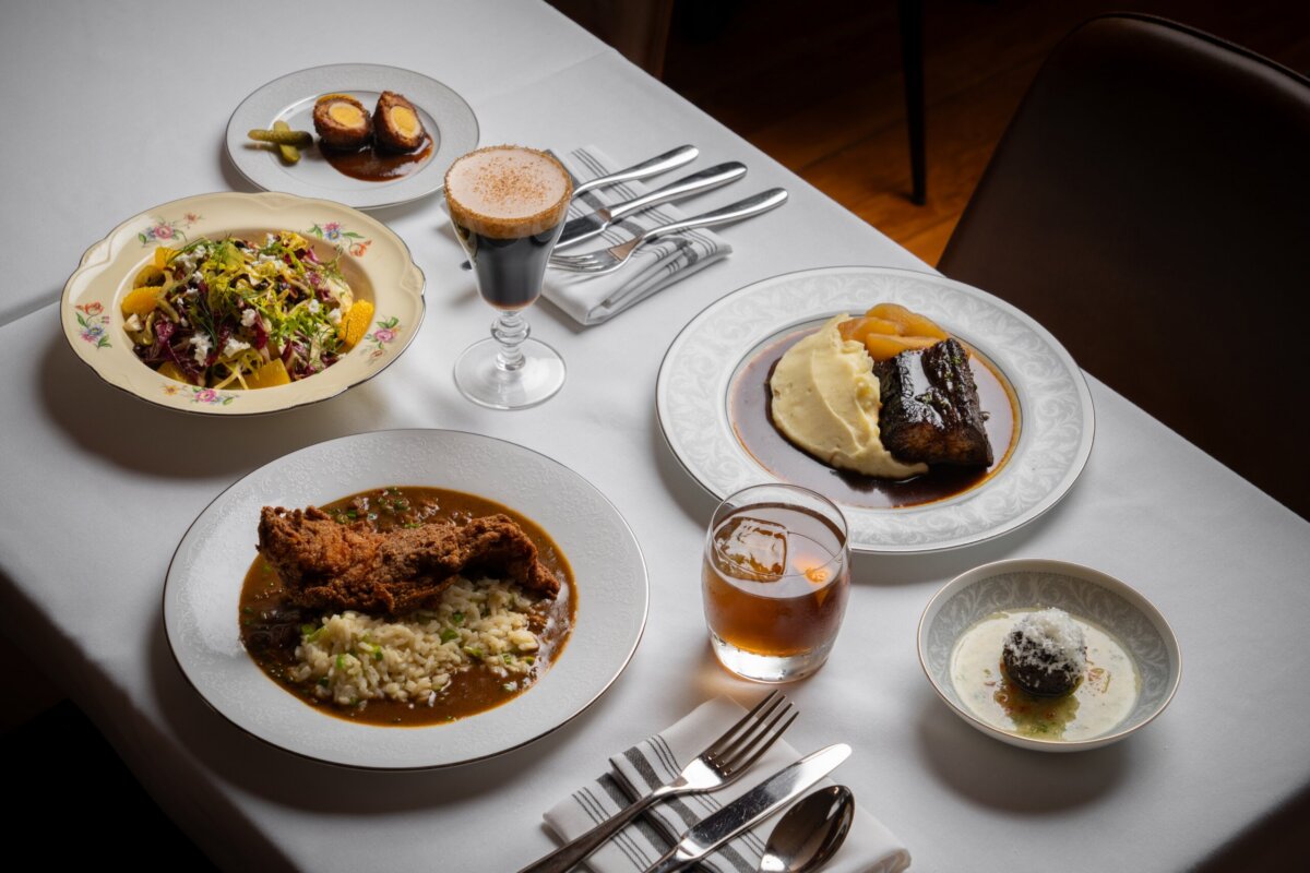 A white tablecloth with five plated dishes, two drinks, and silverware set around them; dishes include salad, fried cutlet, mashed potatoes, dessert, and appetizers.