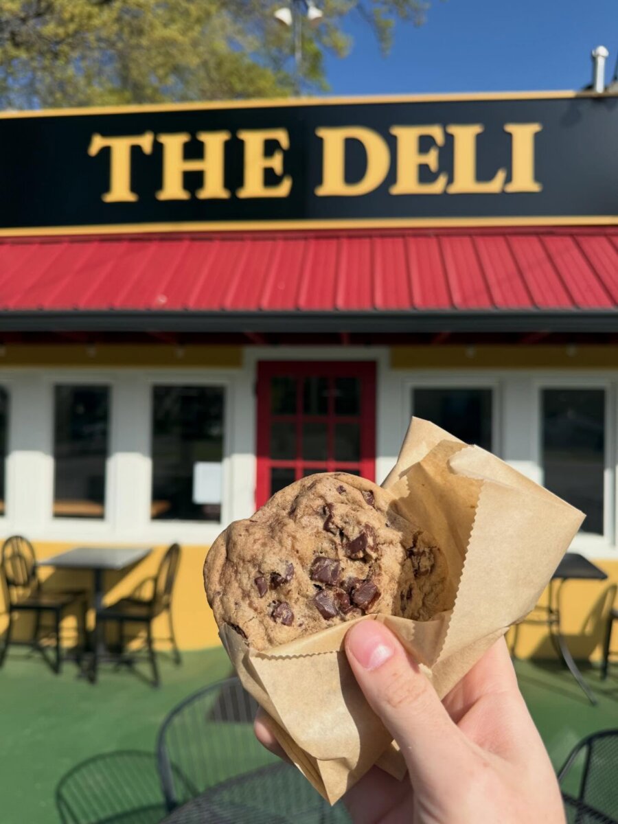 5 New Louisville Restaurants to Try This Month - 2 A hand holds a chocolate chip cookie in a paper sleeve in front of a building with a sign that reads "THE DELI.