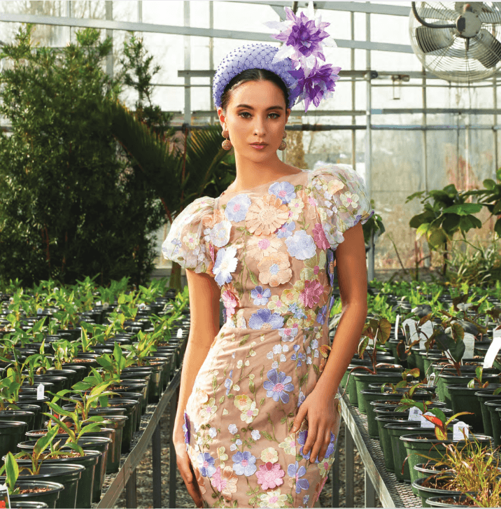 What to Wear for Derby 2026 - 7 Perfect for Derby 2026, this woman showcases Derby fashion in a floral-patterned dress with sheer sleeves and a purple hat adorned with large flowers, standing gracefully among rows of potted plants in a greenhouse.