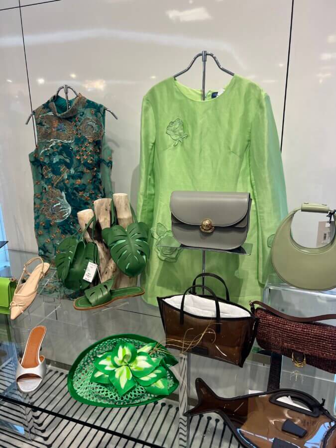 What to Wear for Derby 2026 - 12 Display of women's fashion items—perfect if you're wondering what to wear for Derby fashion—including green dresses, handbags, sandals, and a floral-patterned hat, all arranged on glass shelves against a white wall.