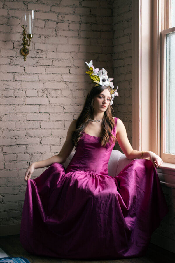 What to Wear for Derby 2026 - 6 A woman in a magenta dress and floral headpiece sits by a window against a brick wall, showcasing what to wear for Derby 2026, with light streaming in from the right.