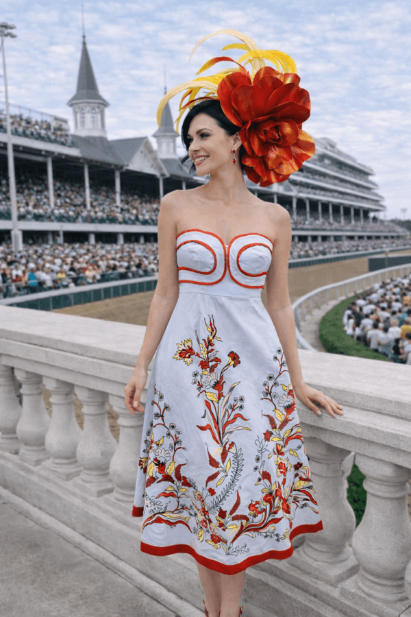 What to Wear for Derby 2026 - 4 A woman in a white dress with red and yellow floral patterns and a bold hat showcases Derby fashion as she stands by a railing at the horse racing stadium, inspiring ideas on what to wear for Derby 2026.