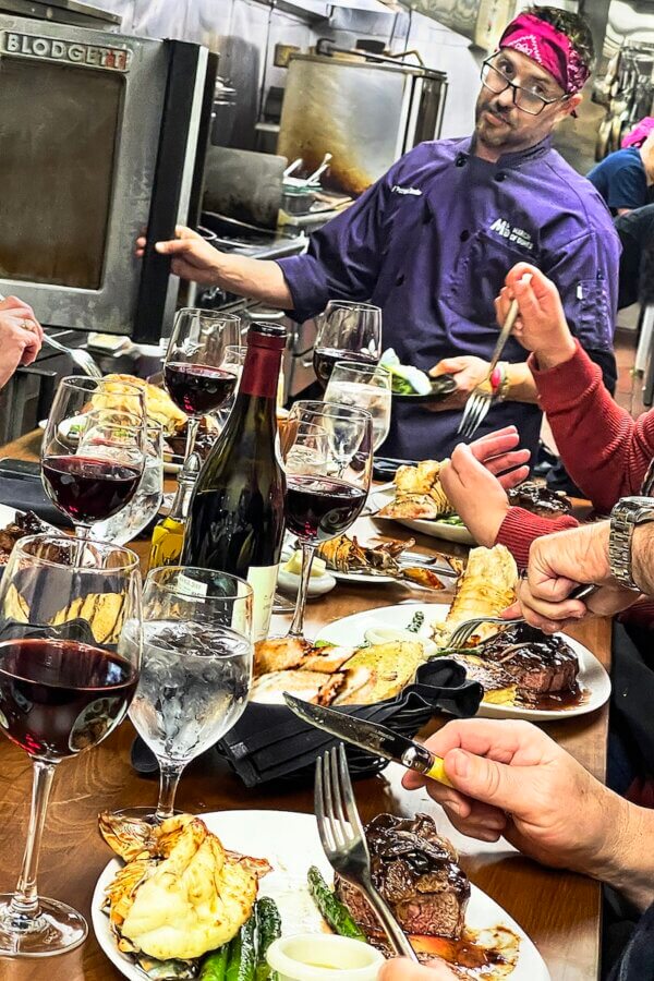 All-Ages Adventure: The Ultimate Trip to Louisville, KY - 9 A chef in a lively Louisville kitchen serves food to diners at a table set with steak, vegetables, and glasses of red wine, creating an inviting adventure for all-ages to enjoy.