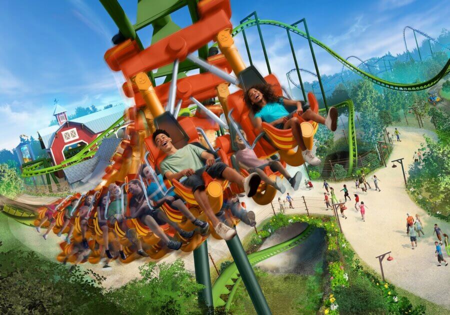 All-Ages Adventure: The Ultimate Trip to Louisville, KY - 7 People enjoy an all-ages adventure on an inverted roller coaster with green tracks at a Louisville amusement park, soaring above a walkway filled with visitors and nearby buildings on a sunny day.