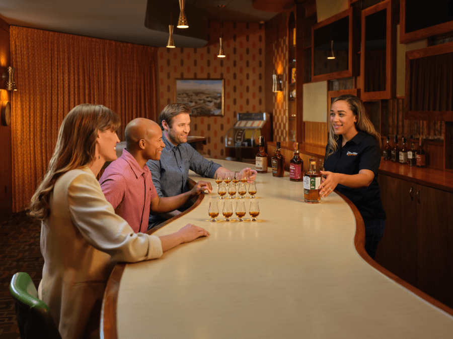 All-Ages Adventure: The Ultimate Trip to Louisville, KY - 15 A bartender serves drinks to three people seated at a curved bar in Louisville, with various liquor bottles lined up on the counter and shelves—an inviting stop on your ultimate trip.